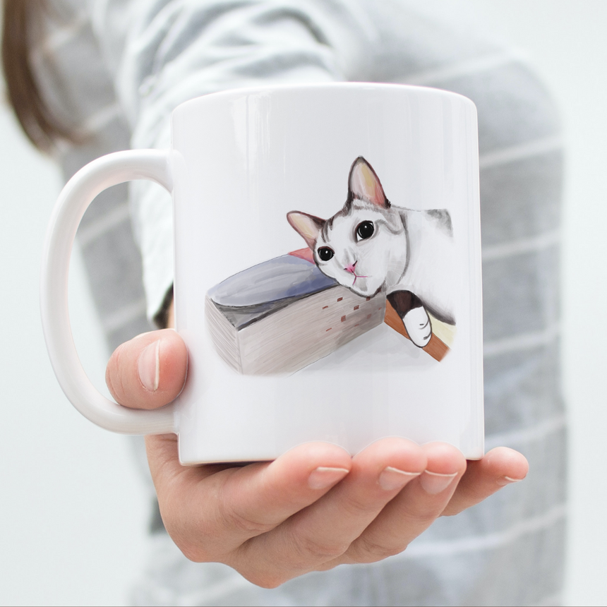 A custom pet portrait mug of a grey cat lying on a book.