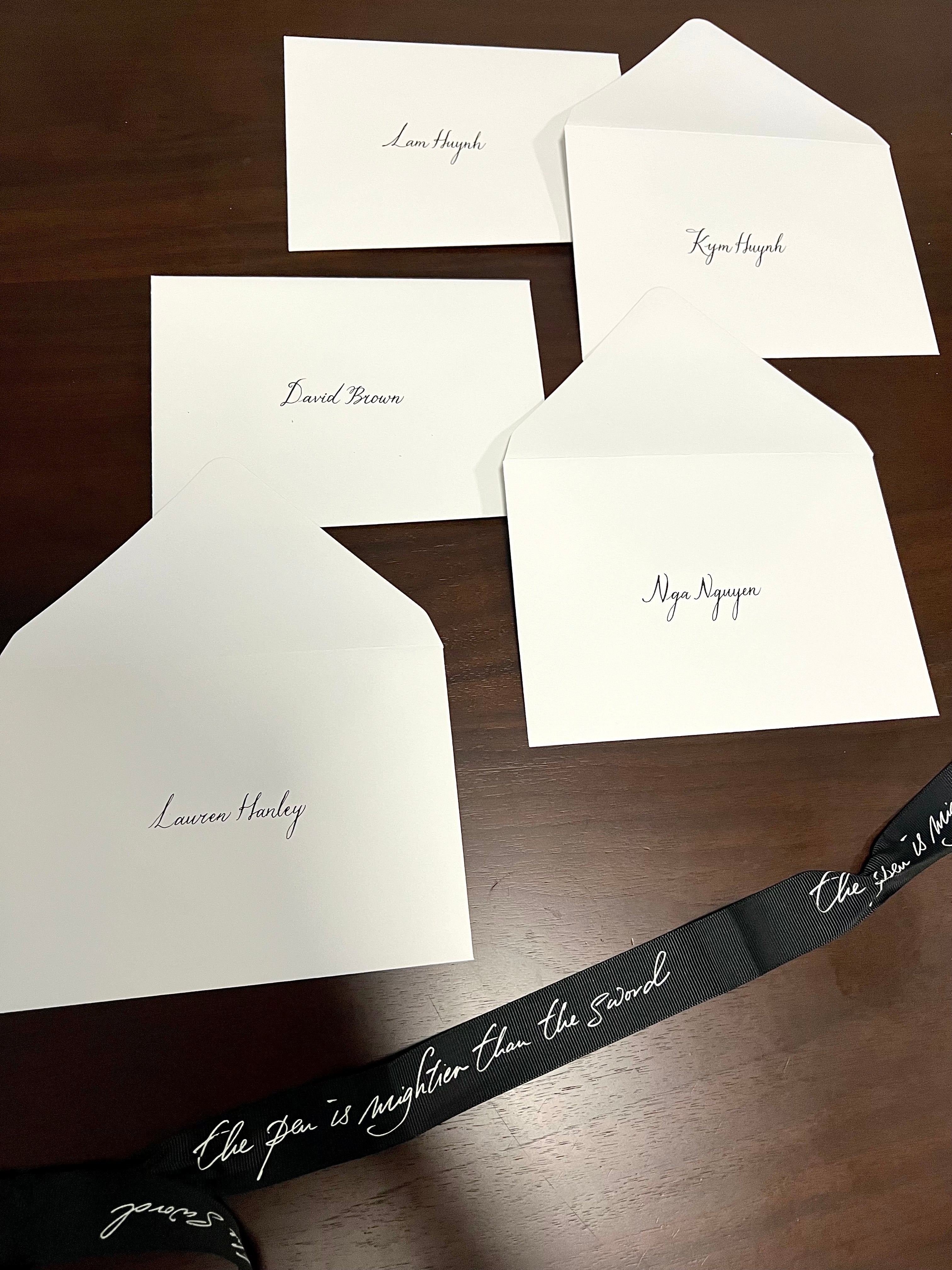 Several envelopes laid out on a surface with handwritten cursive names on them, along with a ribbon with written text.