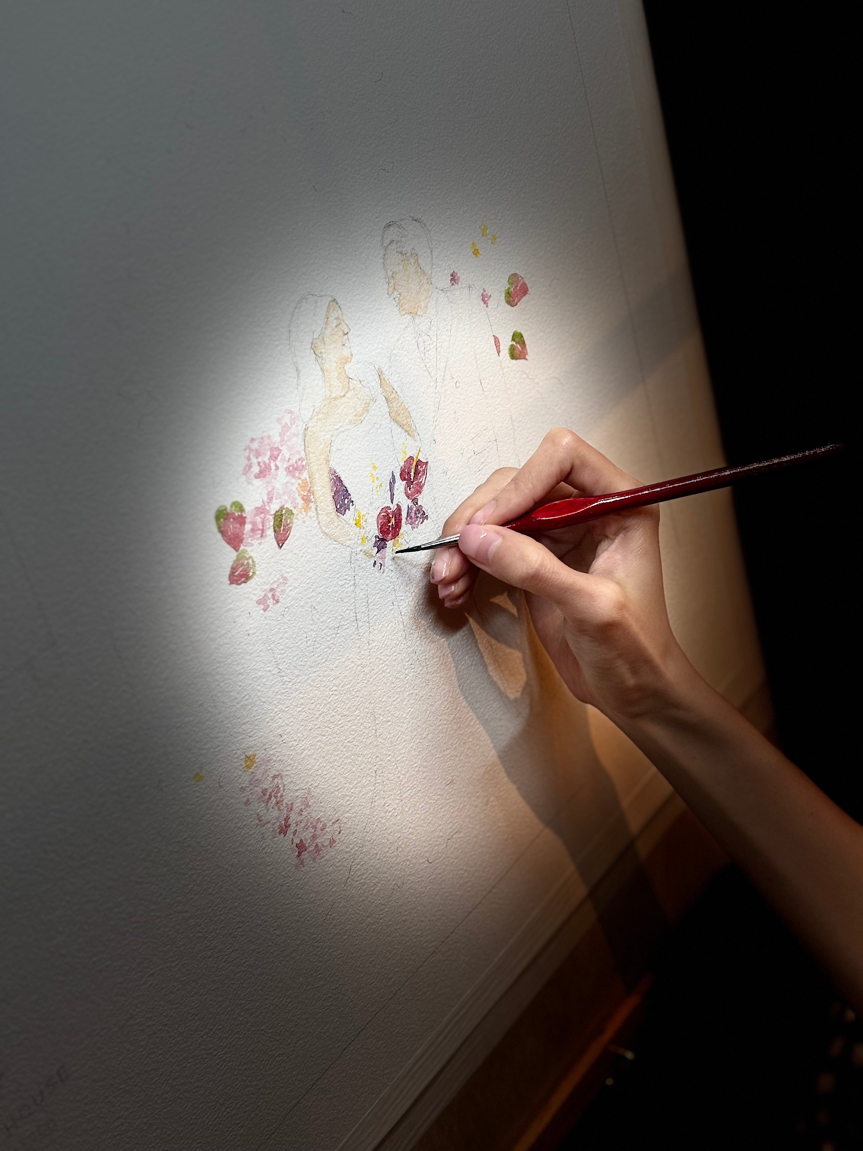 Hand holding a paintbrush over a watercolor painting of flowers on a white surface.
