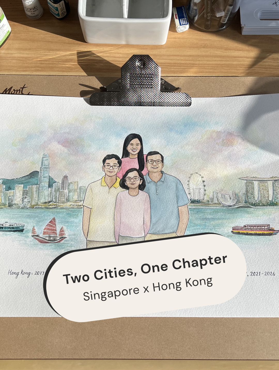 Illustration of a family with a cityscape background and text 'Two Cities, One Chapter: Singapore x Hong Kong' on a wooden surface.