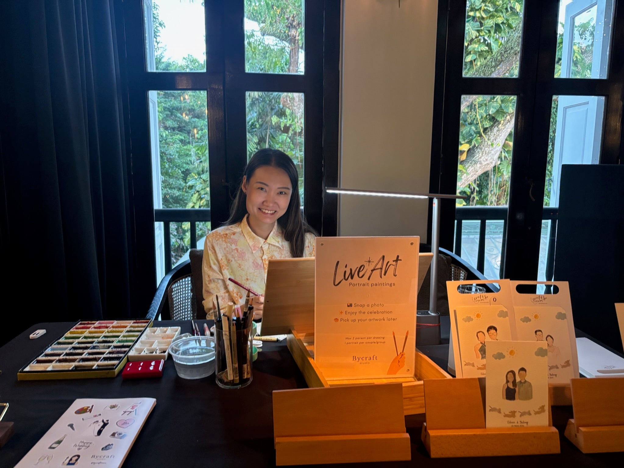 Person sitting at a table with art supplies and promotional materials in an indoor setting.
