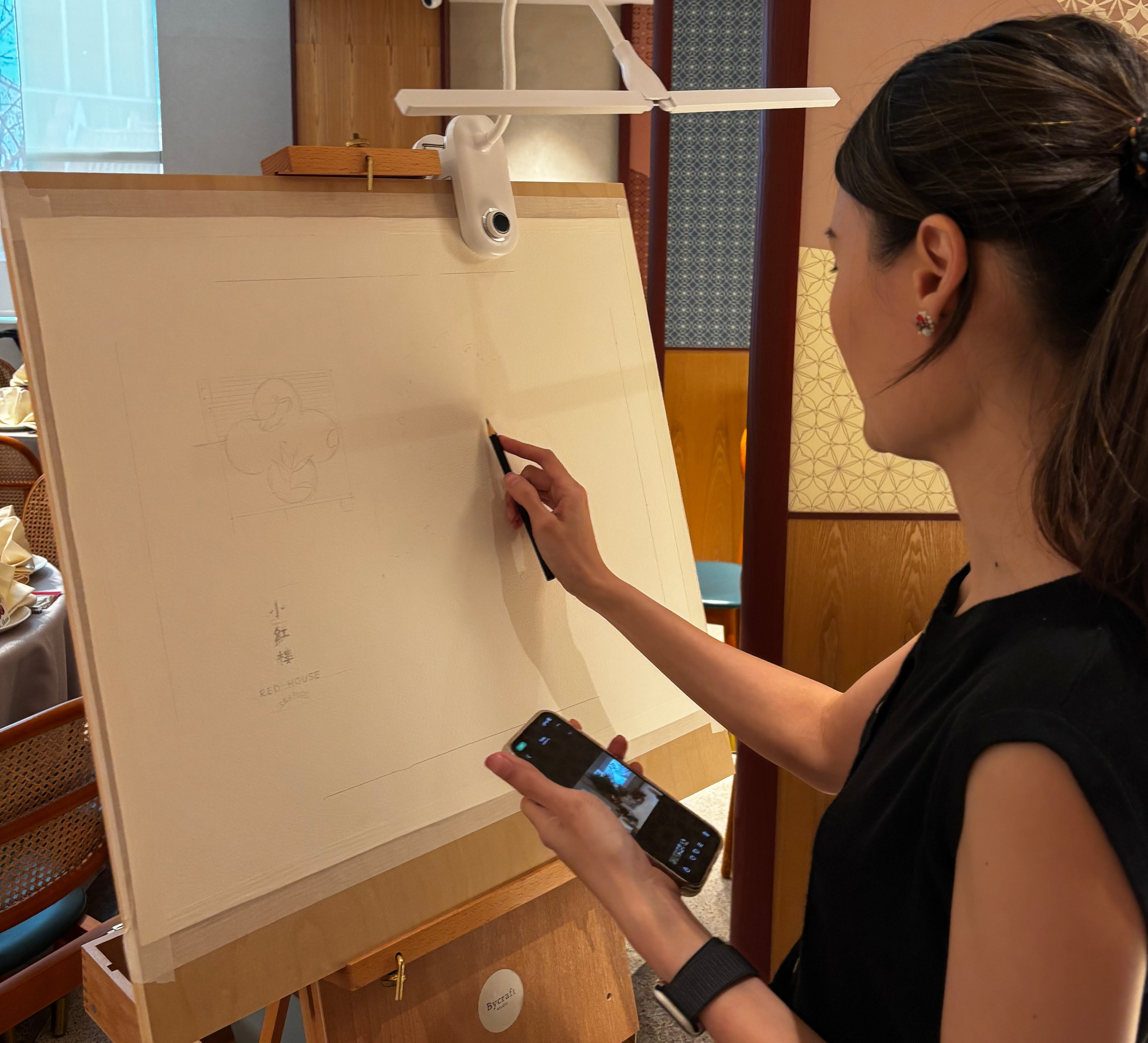 Woman painting on a large canvas with a smartphone for reference in an indoor setting.