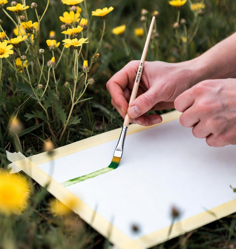 Person painting on a small canvas with a brush, surrounded by yellow flowers.