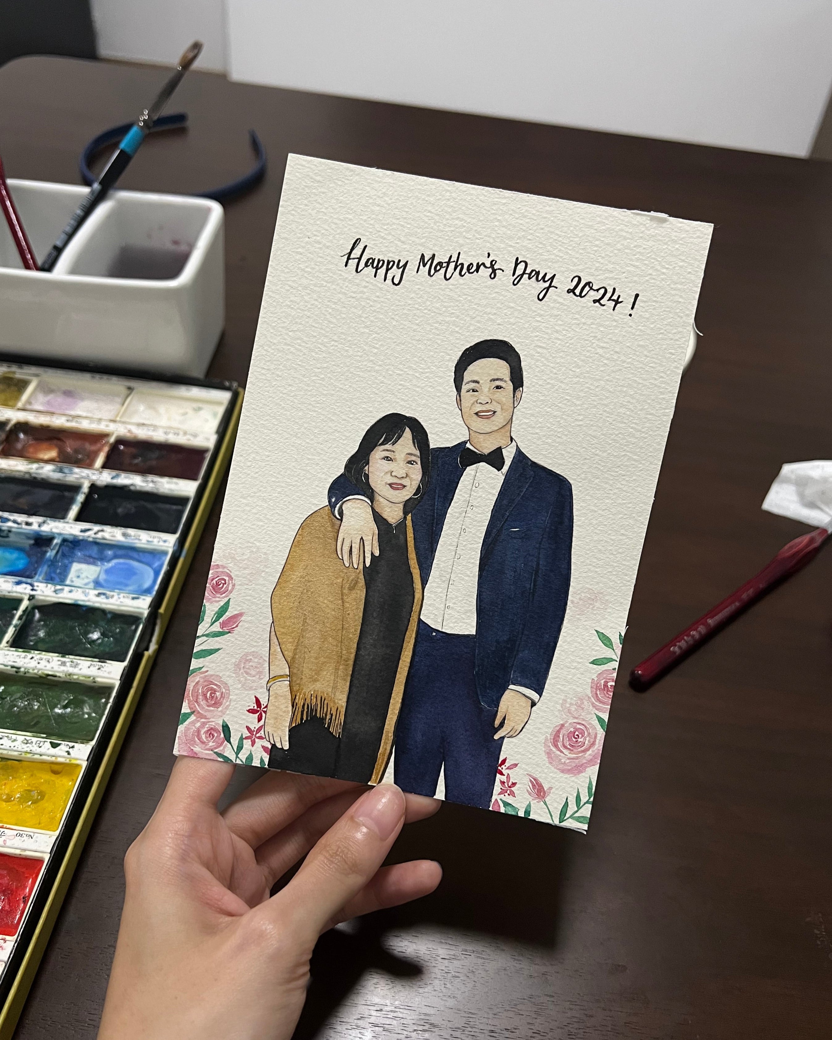 Hand-drawn portrait of a mon and son duo on Mother's Day card with watercolor set and brushes on a table.
