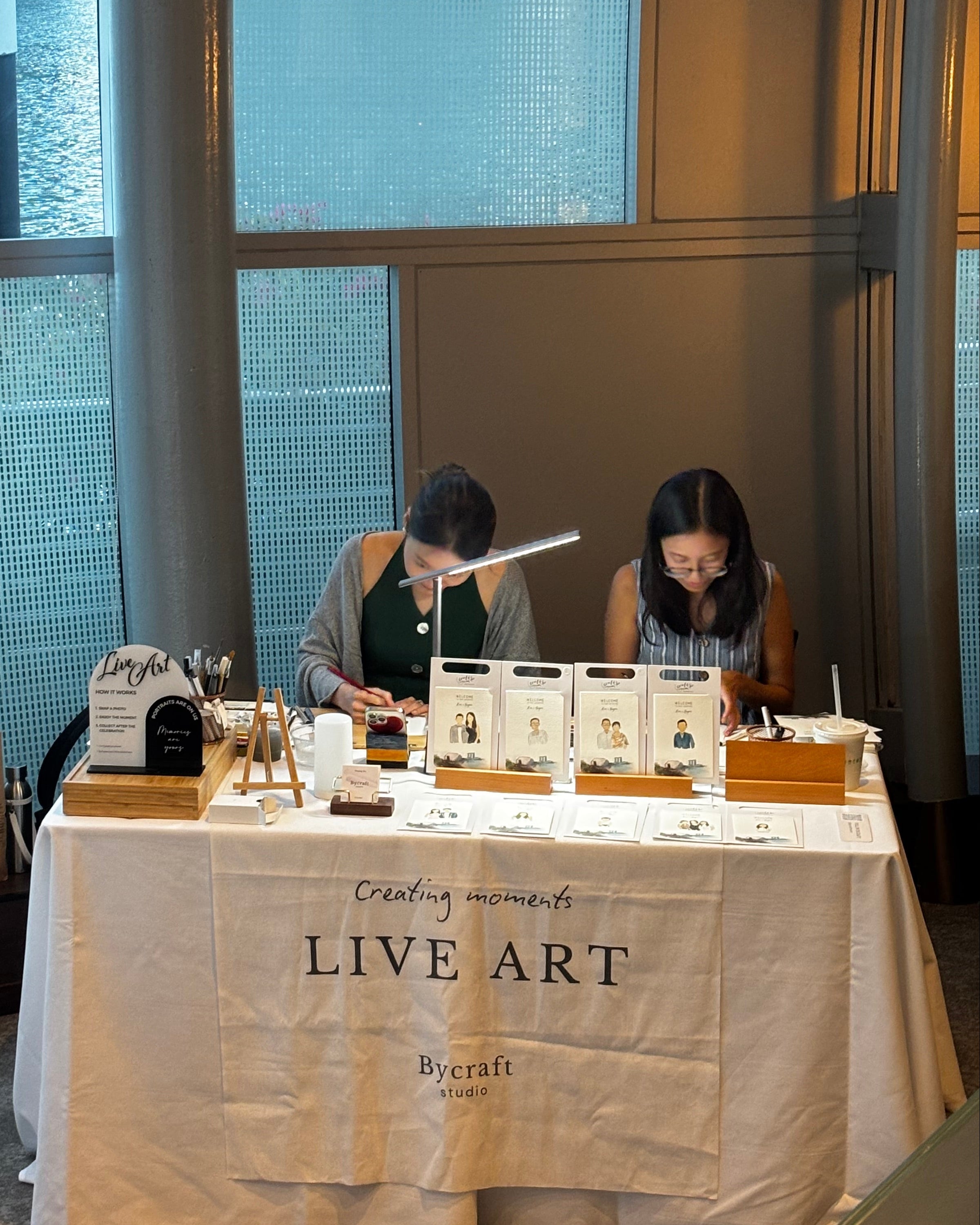Two people standing near a table with a 'Live Art' sign in an indoor setting.