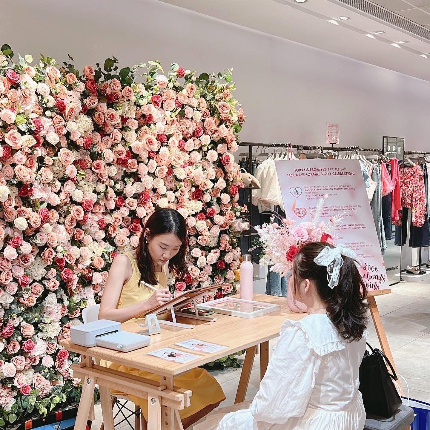 Two women sitting at a table with a floral wall and clothing rack in the background