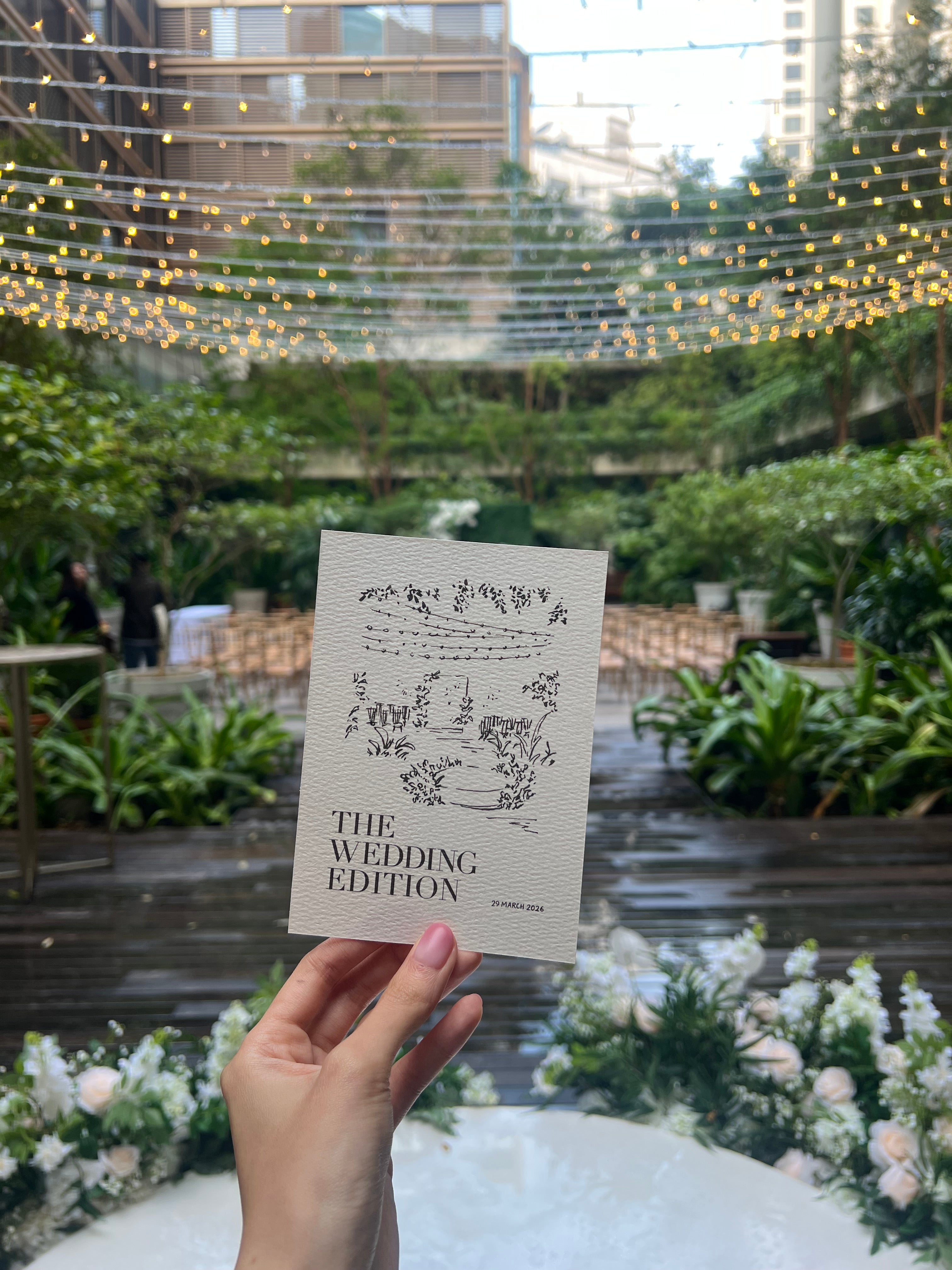 Hand holding a wedding-themed card with decorative background of greenery and flowers.